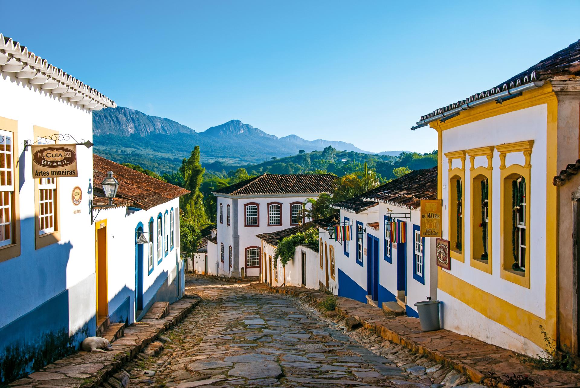 Charmante Kolonialstraße in Tiradentes, Brasilien, mit traditionellen weißen und bunten Gebäuden, Kopfsteinpflasterstraßen und Berglandschaft im Hintergrund an einem sonnigen Tag.